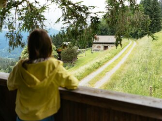 Ein Mädchen genießt von einem Baumhaus aus die Aussicht. Sie schaut in die Ferne, wo man eine Hütte und viele Bäume erkennt. | © Urlaub am Bauernhof Kärnten/ Angelina Tschische