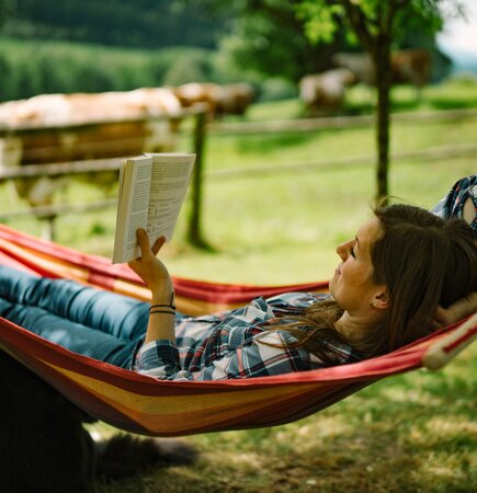 Frau liest Buch in Hängematte | © Urlaub am Bauernhof / Daniel Gollner  Eine Person entspannt in einer bunten Hängematte in einer grünen Landschaft mit Bäumen und Wiesen, liest konzentriert ein Buch. | © Urlaub am Bauernhof / Daniel Gollner