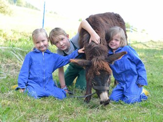 Drei Kinder in Stalloveralls knien neben einem Esel | © Claudia Tschurtschenthaler