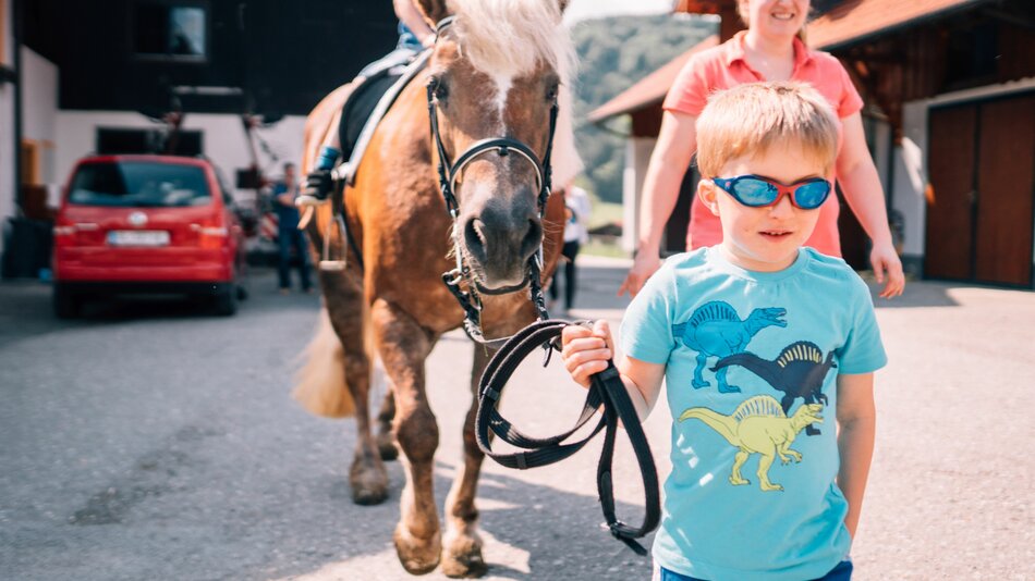 Ein kleiner Bub führt ein Pferd mit Reiter über das Hofgelände. | © Urlaub am Bauernhof im SalzburgerLand / Daniel Gollner