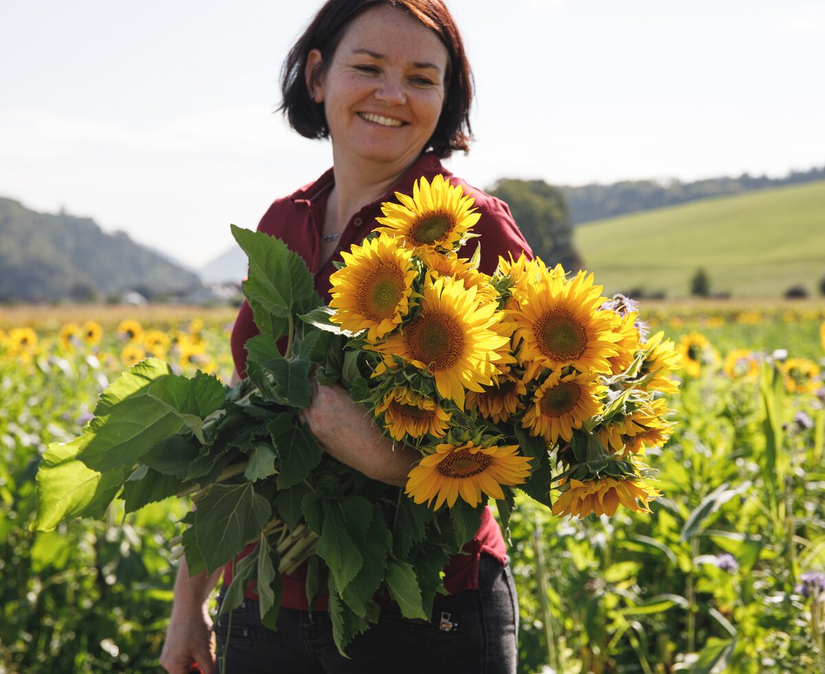 Bäuerin steht mit einem Strauß gepflückter Sonnenblumen im Feld | © Urlaub am Bauernhof / Punkt & Komma